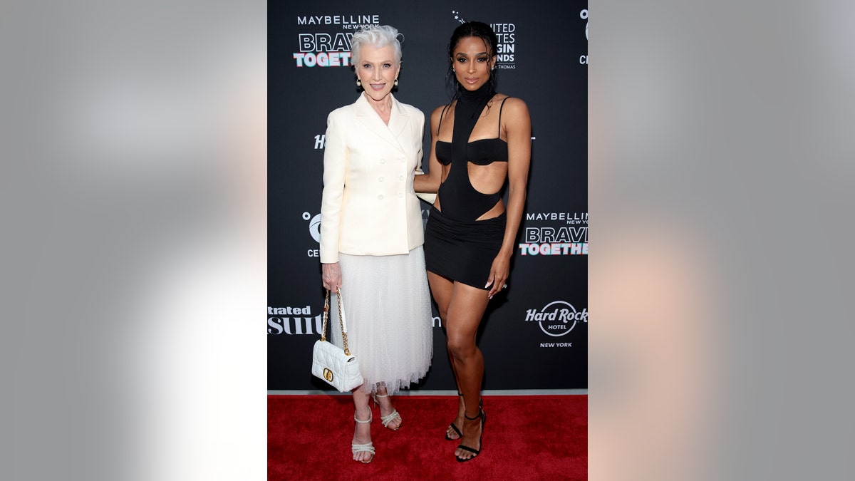 Maye Musk wearing a white blazer and a white ballerina skirt posing next to Ciara who is wearing a black cut-out dress