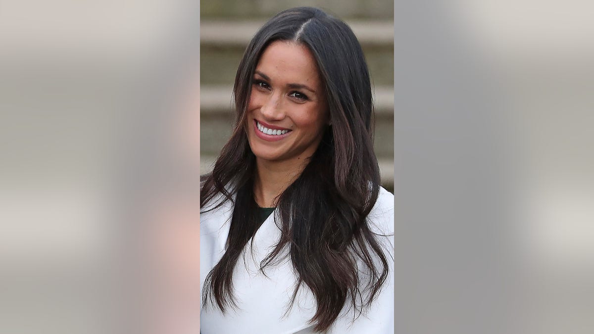 A close-up of Meghan Markle smiling and wearing a white blazer with a black shirt.