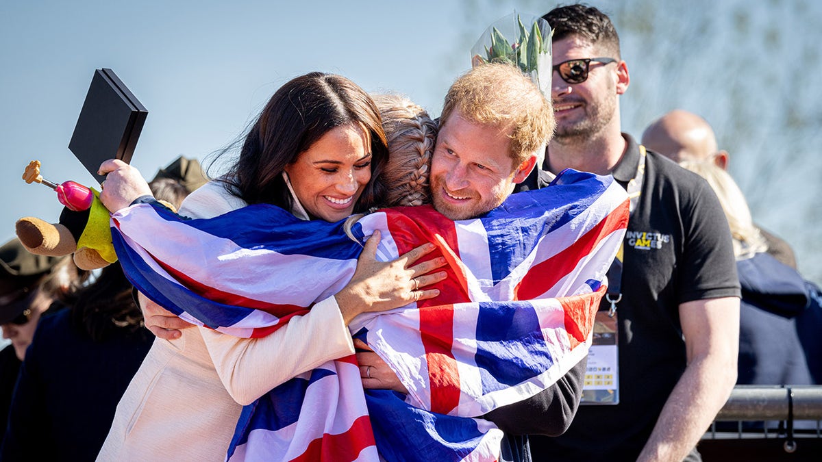 Prince Harry and Meghan Markle embracing someone with the Union Jack flag around them.