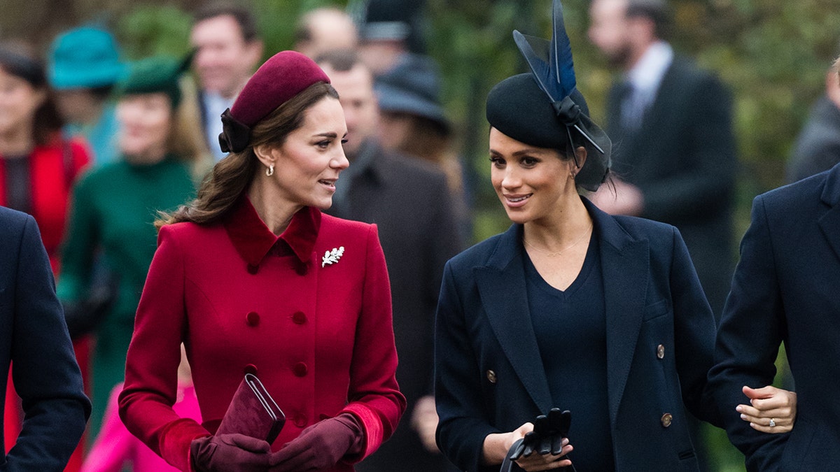 Kate Middleton in a red coat chatting with Meghan Markle in a dark blue coat.