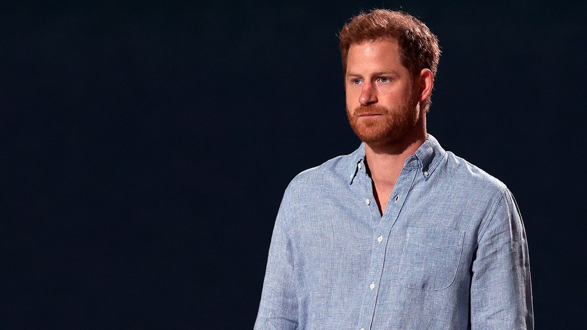 Prince Harry looking serious in a light blue shirt against a black background.