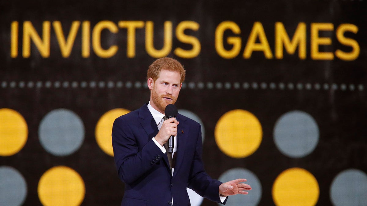 Prince Harry talking to a mic in a navy suit with the Invictus Games logo behind him