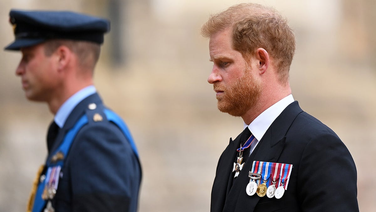 Prince Harry looking somber in a suit with medals walking behind Prince William in uniform.