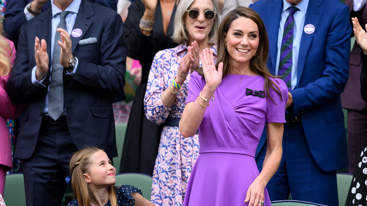 Kate Middleton waving in a purple dress as Princess Charlotte looks up at her from the stands.