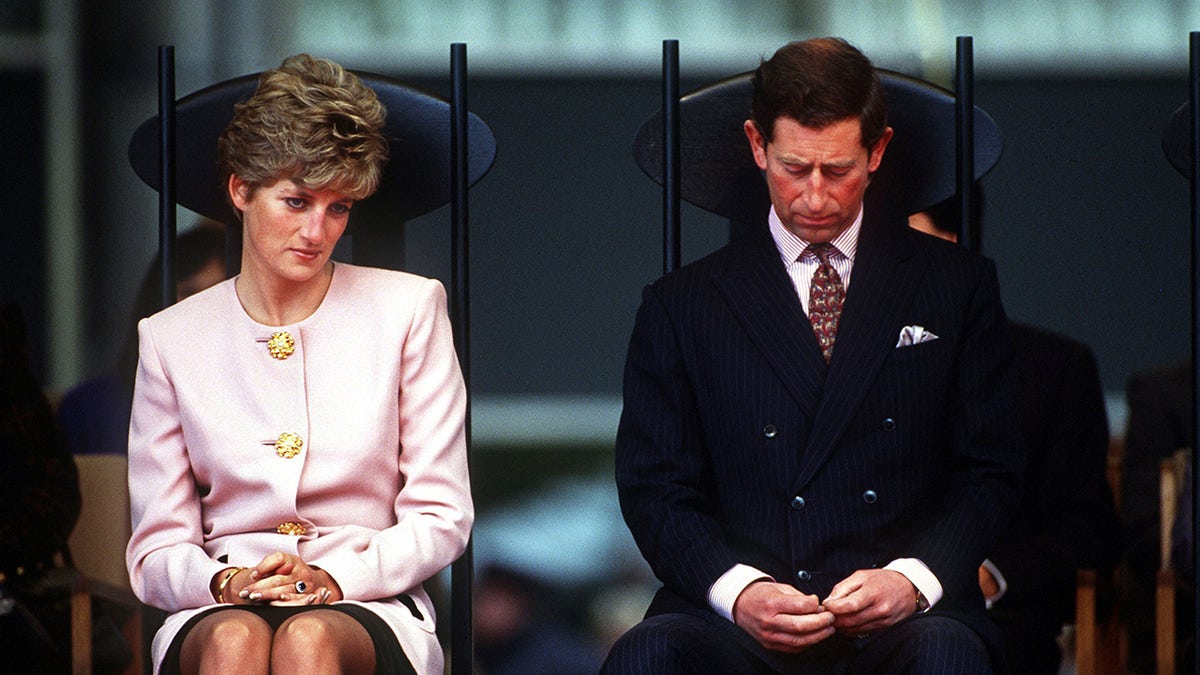 Diana in a pink suit dress looking down and sitting next to Prince Charles in a dark suit.