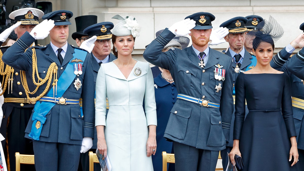 Prince Harry and Prince William saluting in blue uniforms as their wives look serious ahead.