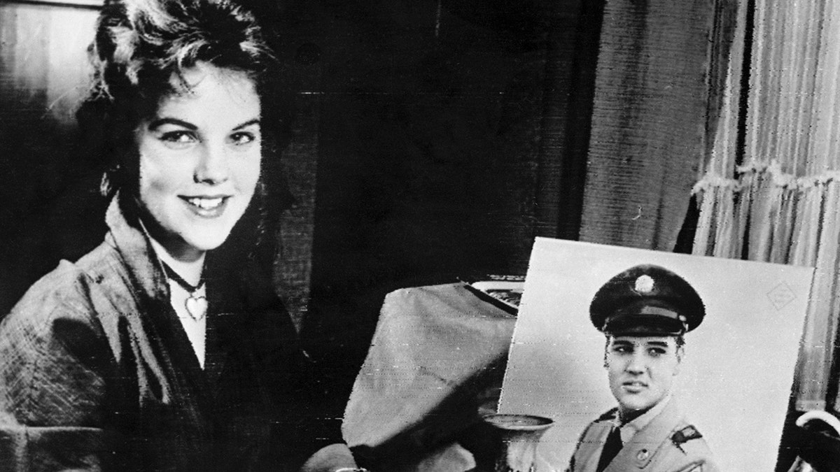 Priscilla Presley smiling next to an Army portrait of Elvis Presley