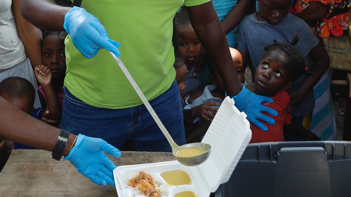 A server wearing blue rubber gloves ladles soup into a container as child in line for food looks up.