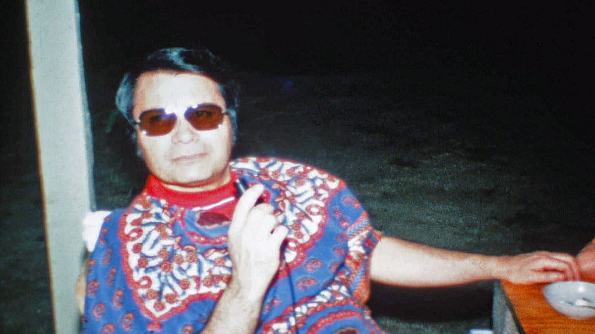 Jim Jones wearing a colorful sweater holding a mic.