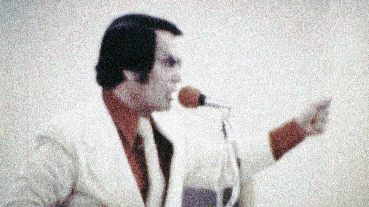 Jim Jones looking angry in a red shirt and a white blazer and a matching tie preaching to a red mic.