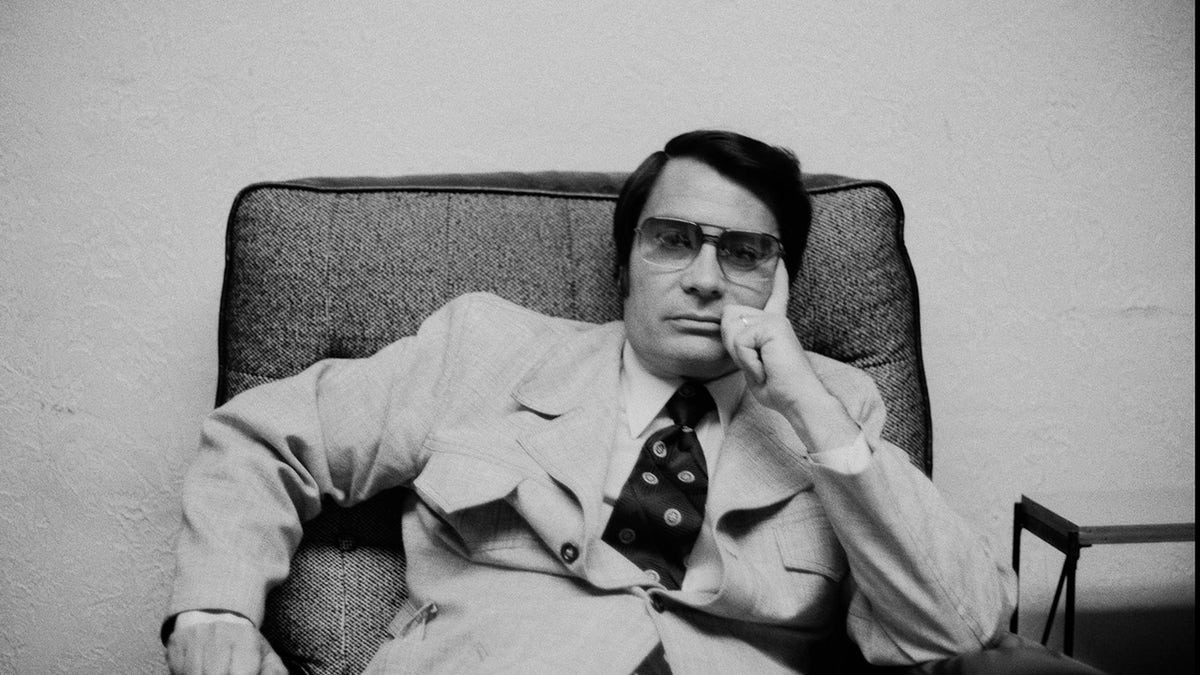Jim Jones in a suit and tie sitting on a couch dazed.