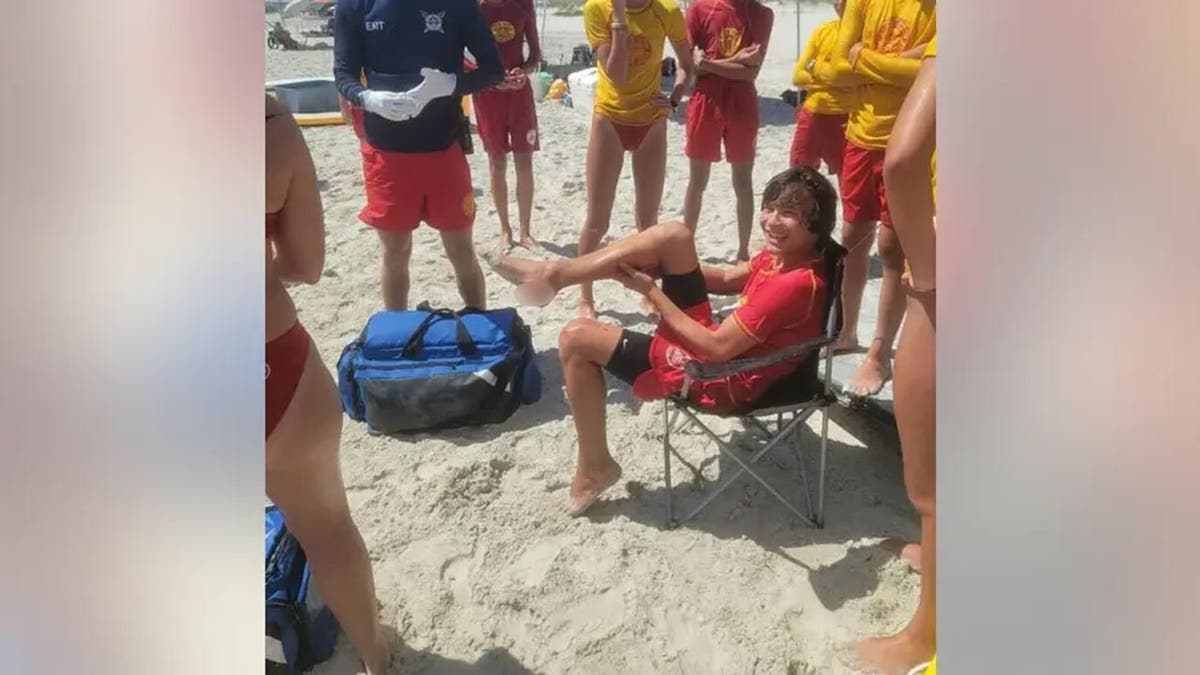Boy sits on a chair smiling while holding his leg up high after being bitten by a shark