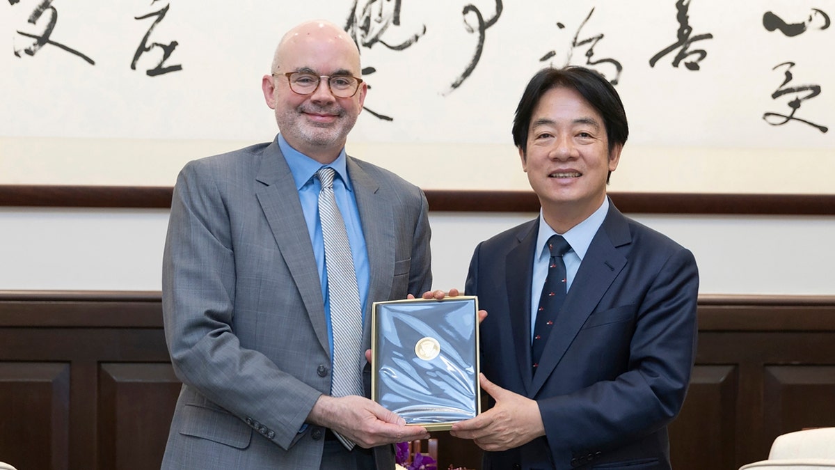 Taiwan's President William Lai Ching-te, right, poses for photos with American Institute in Taiwan's director Raymond F. Greene, left. Both appear in suits and are holding a plaque between them.