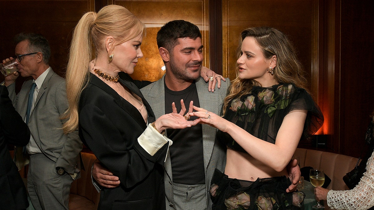 Nicole Kidman con un blazer negro habla con Joey King con un conjunto de dos piezas negro de encaje mientras ambas rodean con los brazos a Zac Efron con camisa negra y traje gris