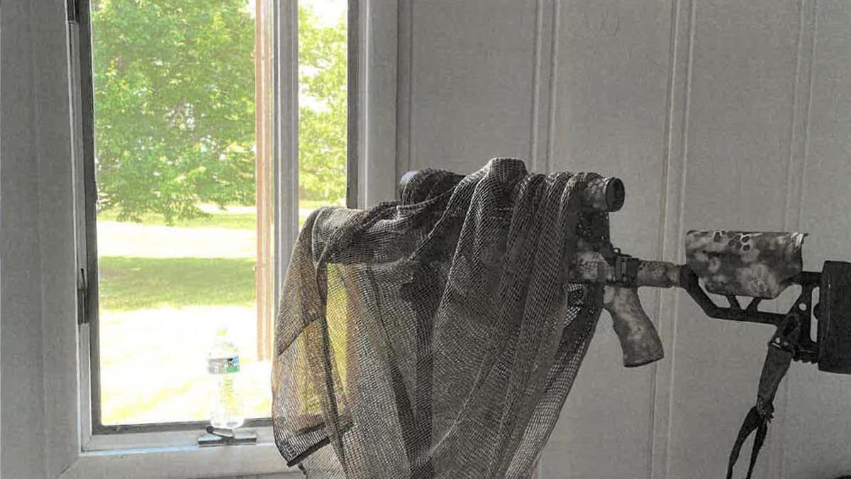 A sniper's rifle is camouflaged in gauze as it sits on a stand, pointing out the window