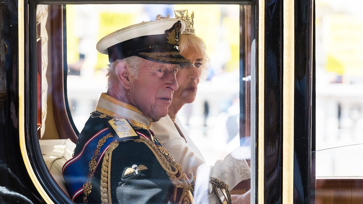 King Charles wearing a royal uniform sitting in a glass carriage next to Queen Camilla.