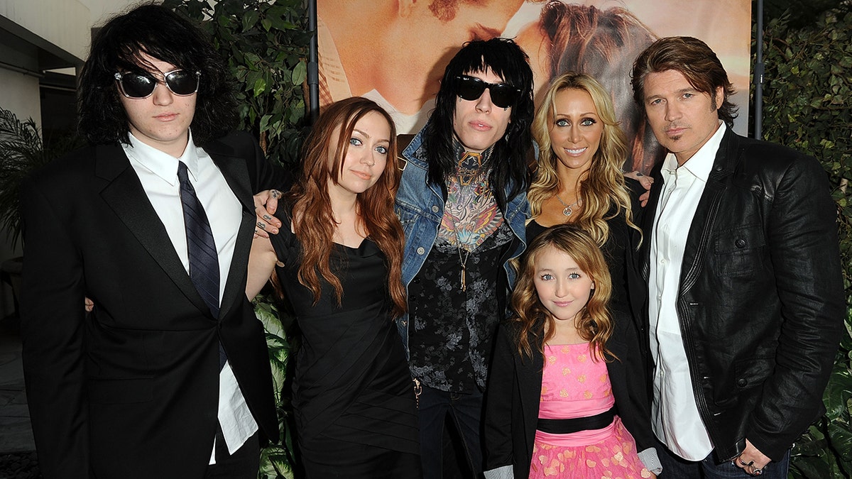 Braison Cyrus in a black suit, next to Brandi Cyrus in a black dress next to Trace Cyrus in a jean jacket next to Tish and Billy Ray and beneath them is Noah in pink