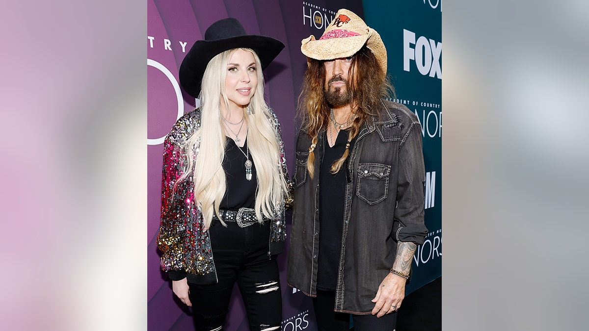 Firerose in a black hat and long blonde hair poses next o Billy Ray in a dark grey shirt and straw hat
