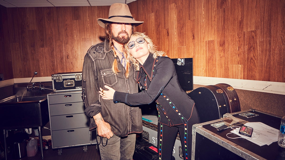 Billy Ray Cyrus in a brown shirt and hat is hugged by daughter Miley Cyrus in black