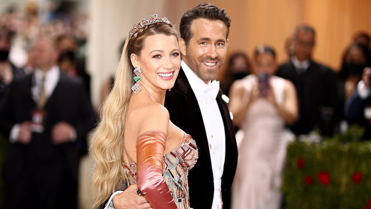 Blake Lively turns back at the Met Gala in a multi-colored peach dress and tiara with Ryan Reynolds in a tuxedo smiling at the camera