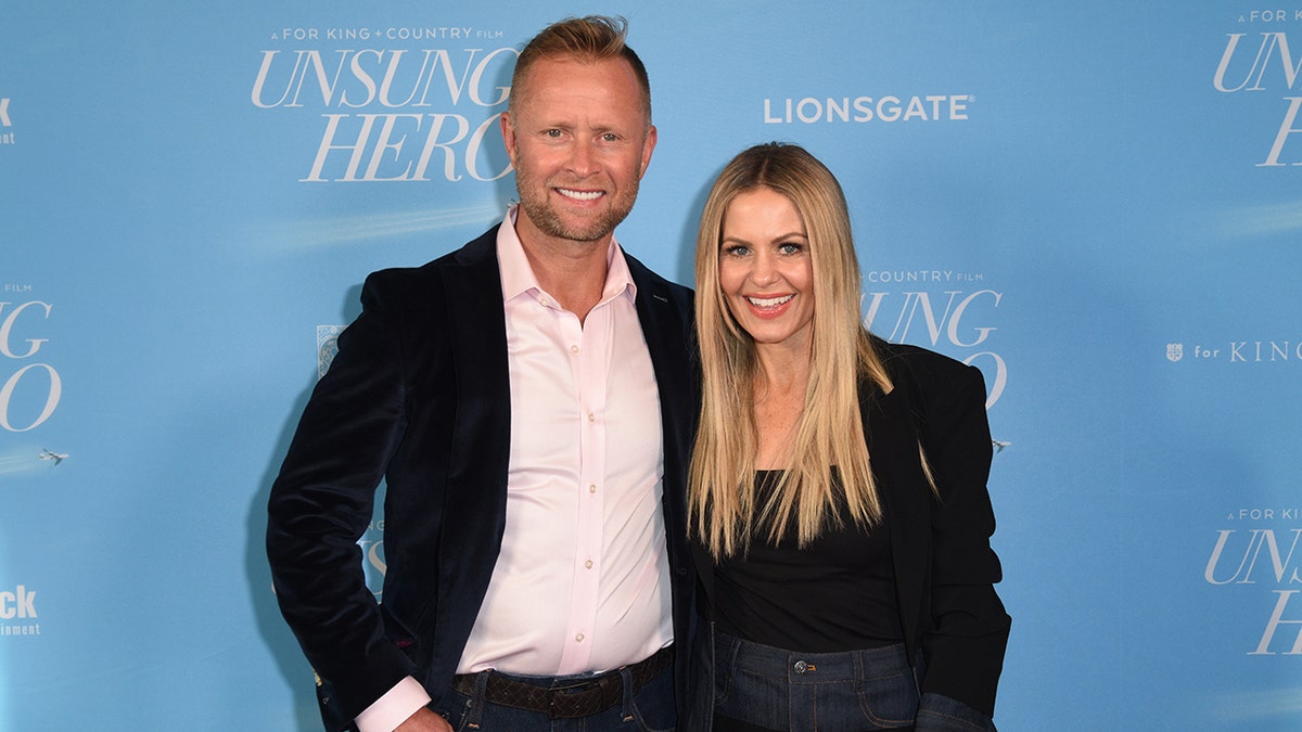 Valeri Bure in a black suit and light pink shirt stands on the carpet next to wife Candace Cameron Bure in a black outfit