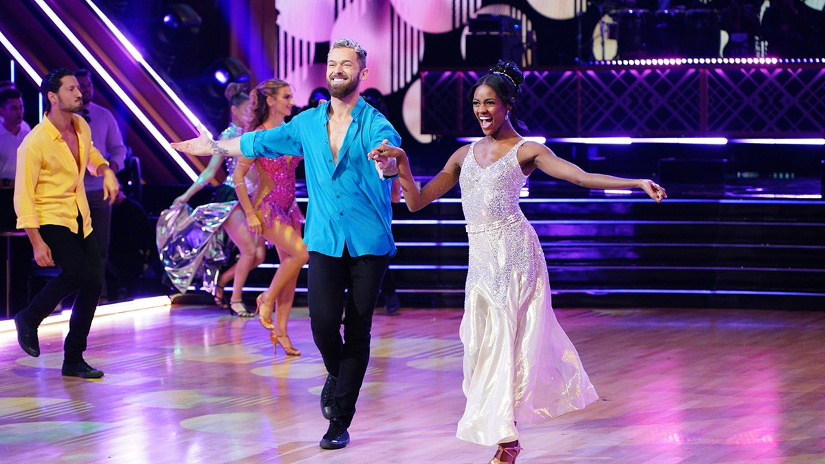 Artem Chigvintsev in a blue shirt and black pants dances with Charity Lawson in a long white dress