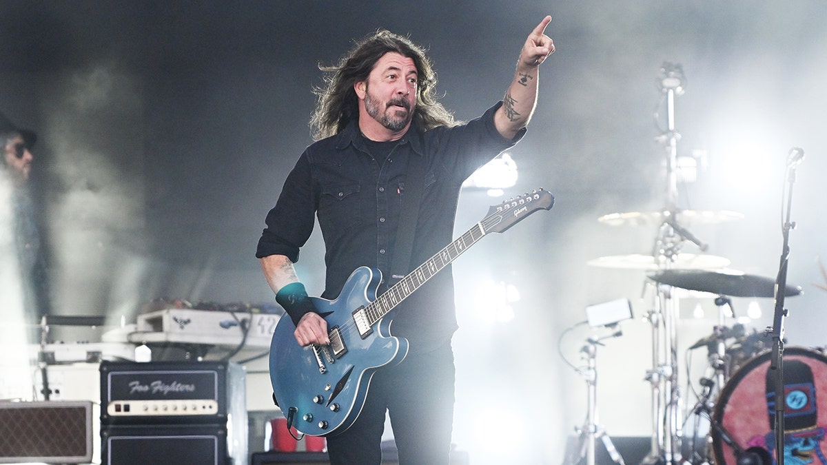 Dave Grohl in a black shirt points his finger out towards the audience while on stage with a blue guitar