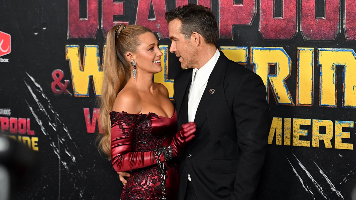 Blake Lively in a red printed jumpsuit holds onto husband Ryan Reynolds in a black suit and white shirt