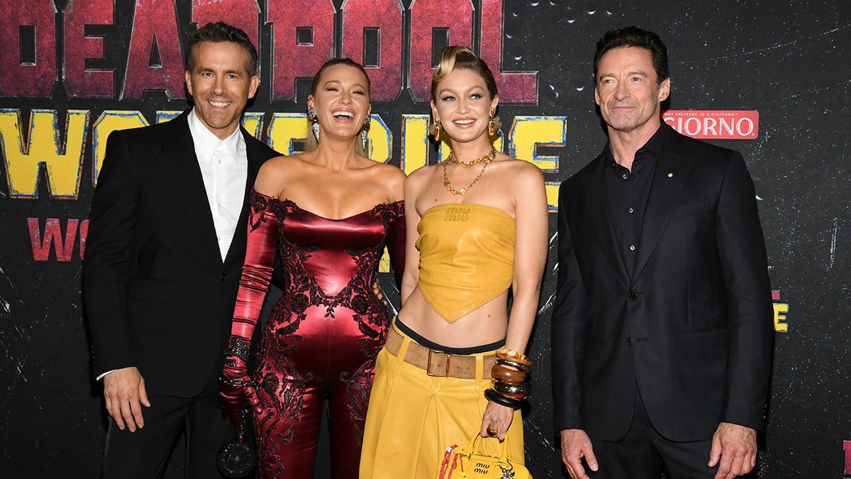 Ryan Reynolds in a black suit and white shirt laughs next to wife Blake Lively in a red metallic jumpsuit next to Gigi Hadid in a yellow two piece outfit next to Hugh Jackman in a black suit and shirt at the premiere of "Deadpool & Wolverine"