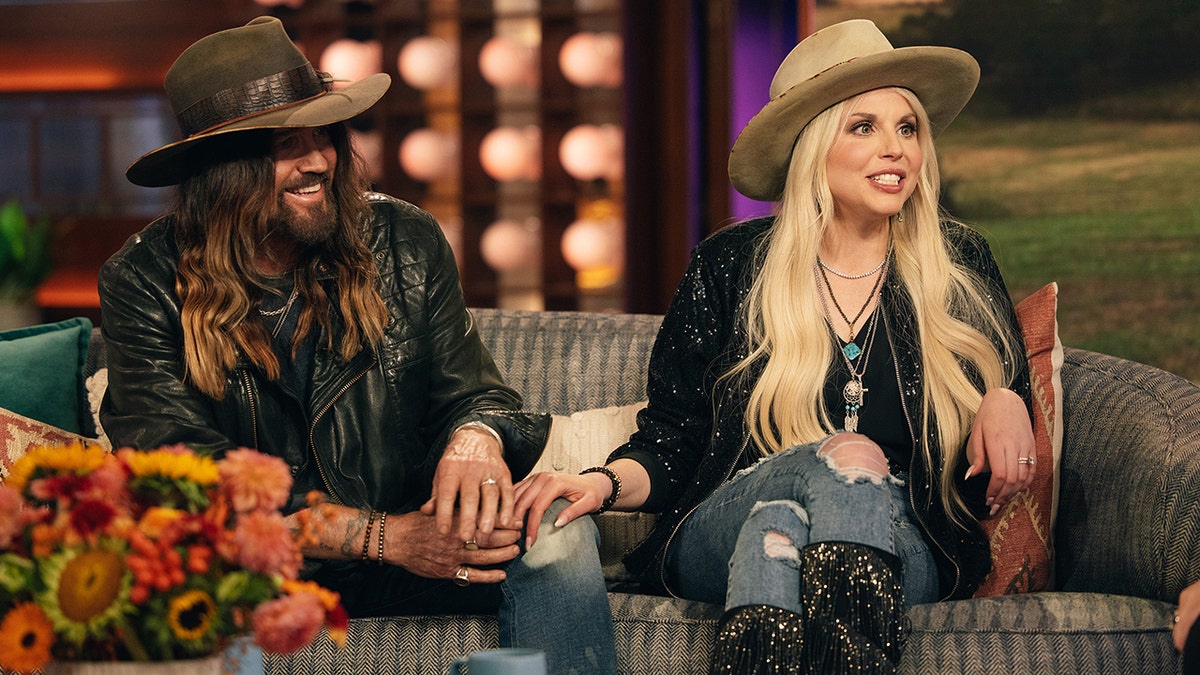 Billy Ray Cyrus on the couch holding a guitar is seated next to wife Firerose on "The Kelly Clarkson Show"
