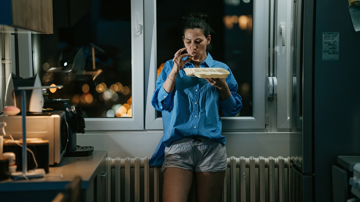 Mulher em pé na cozinha comendo tarde da noite