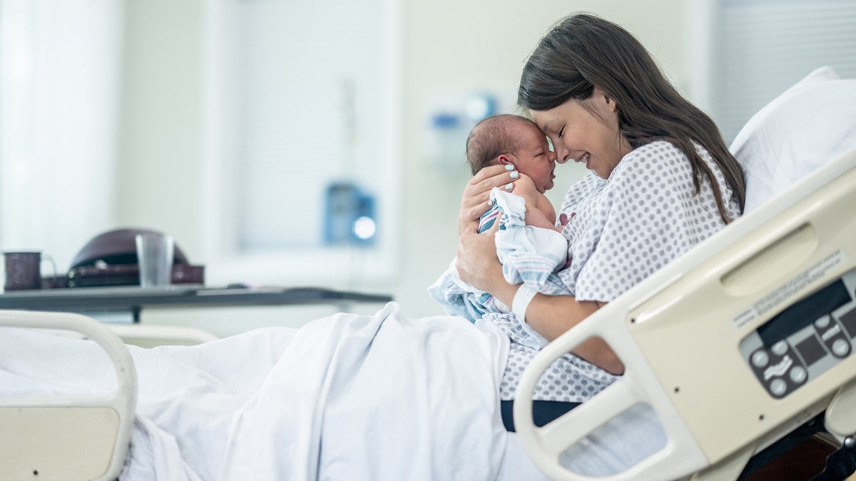 A new Mother sits up in her hospital bed shortly after delivery as she holds her newborn out in front of her