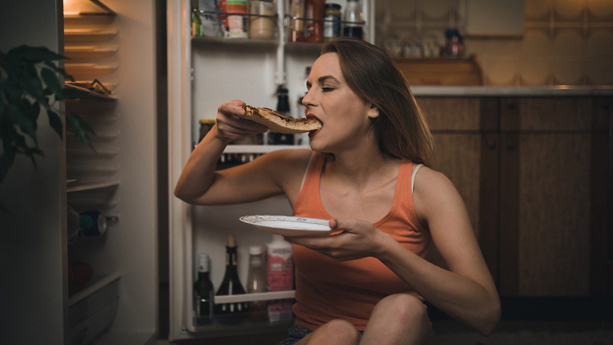 Woman eating pizza slice in front of the refrigerator late night