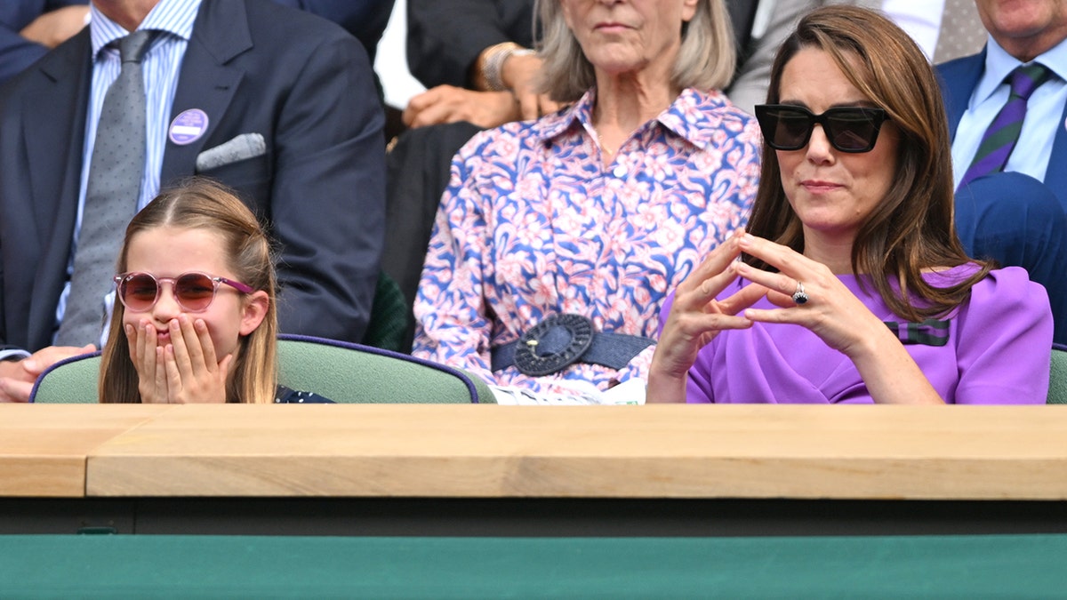 Kate Middleton in a purple dress and black sunglasses puts her fingers together as she watches Wimbledon next to Princess Charlotte who covers her mouth