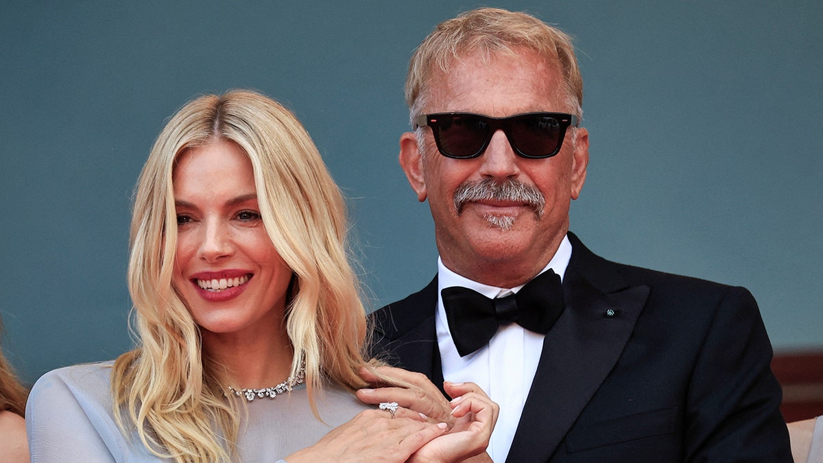 Sienna Miller in a periwinkle dress holds hands with Kevin Costner in a tuxedo and black glasses