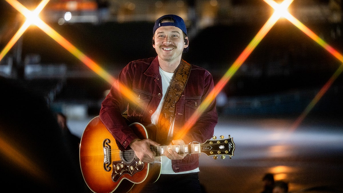 Morgan Wallen con una chaqueta roja tocando la guitarra con un sombrero al revés