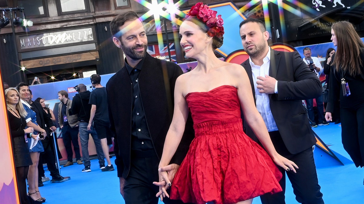 Benjamin Millepied in black holds hands with Natalie Portman in a red dress and floral hair piece