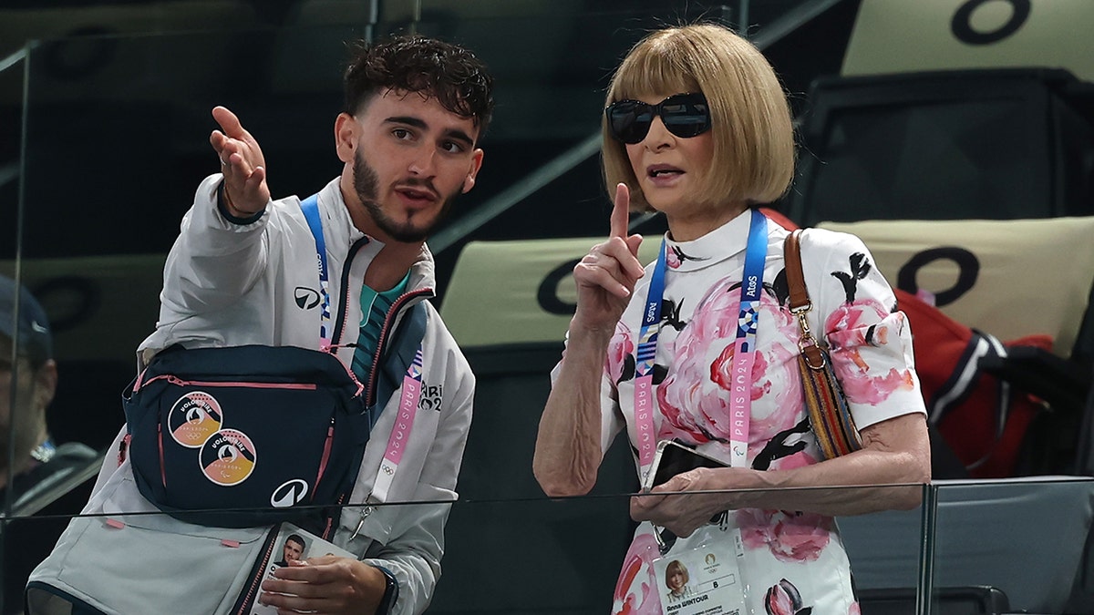 Anna Wintour in a colorful dress points outward next to a man at the Olympics