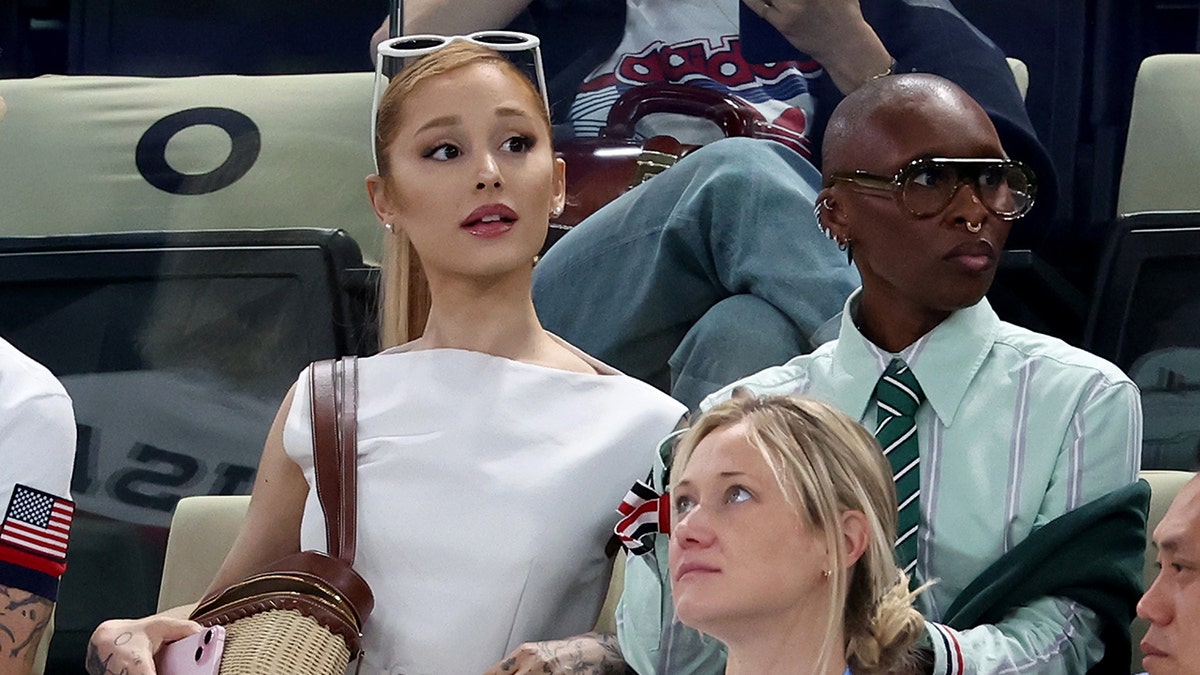 Ariana Grande in a white dress looks up seated next to Cynthia Erivo in a mint green striped shirt