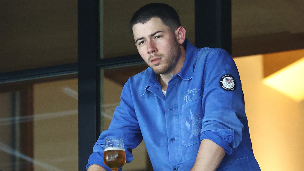 Nick Jonas in a blue zip up looks down at the Olympics