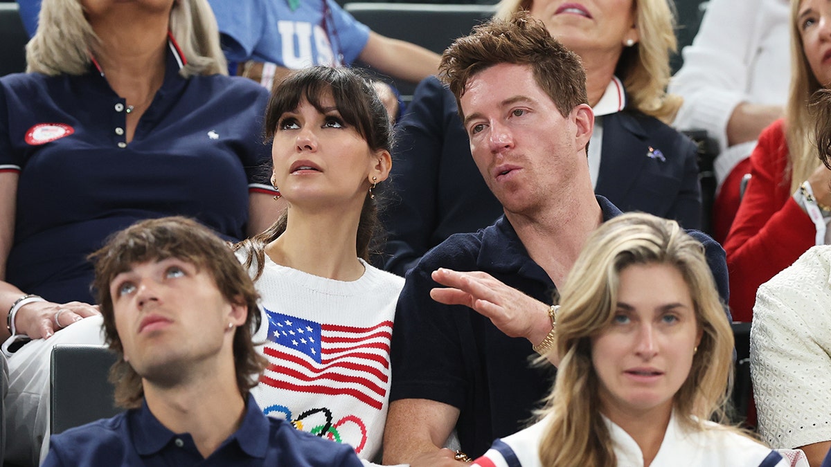 Nina Dobrev in a white USA Olympics sweatshirt looks up as she is seated next to boyfriend Shaun White at the Olympics
