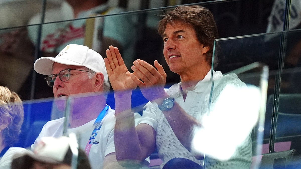 Tom Cruise sitting behind glass caps his hands watching the Artistic Gymnastics