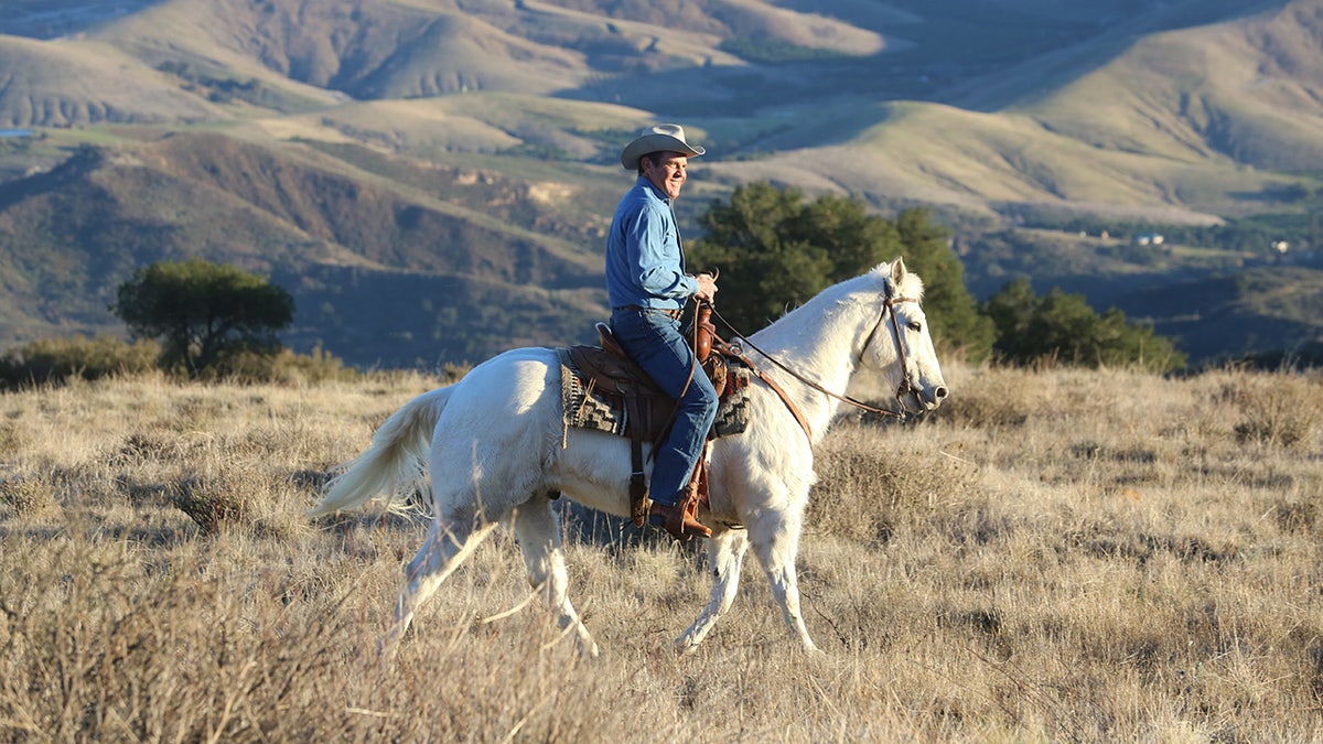 Dennis Quaid wears a cowboy hat while riding a horse in the hills.