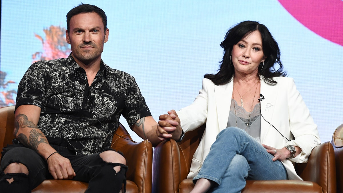Brian Austin Green in a patterned shirt sits in a brown chair and holds Shannen Doherty's hand in a white blazer and jeans