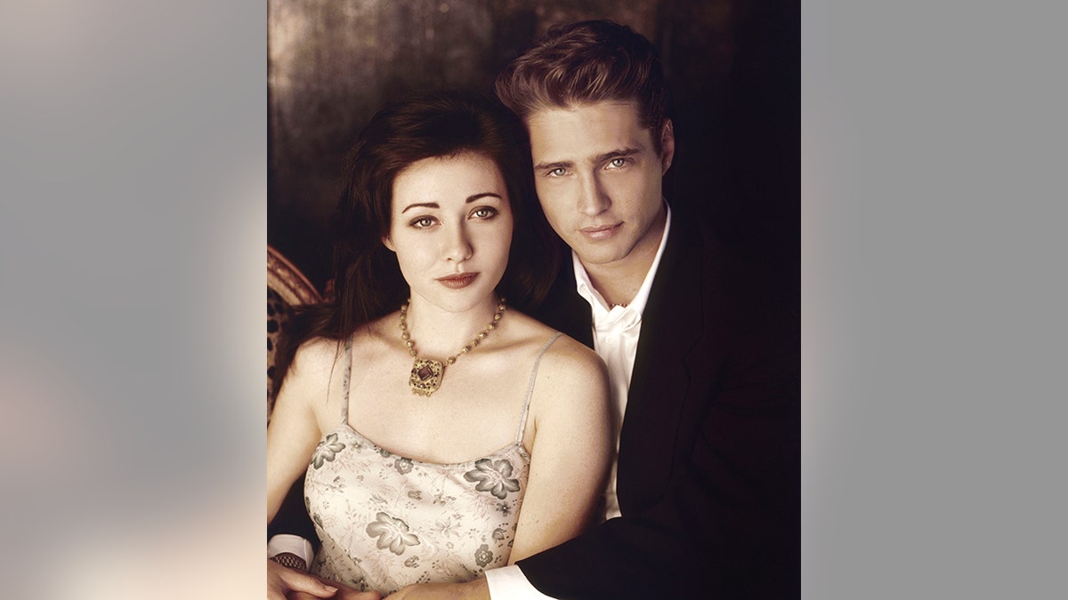 Light colored photo of Shannen Doherty in a tank top being held by Jason Priestley in a black suit