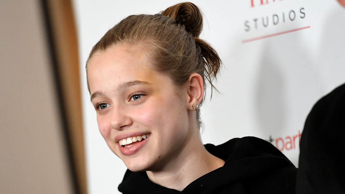 Shiloh Jolie in a black sweatshirt smiles with her hair in a bun on the carpet