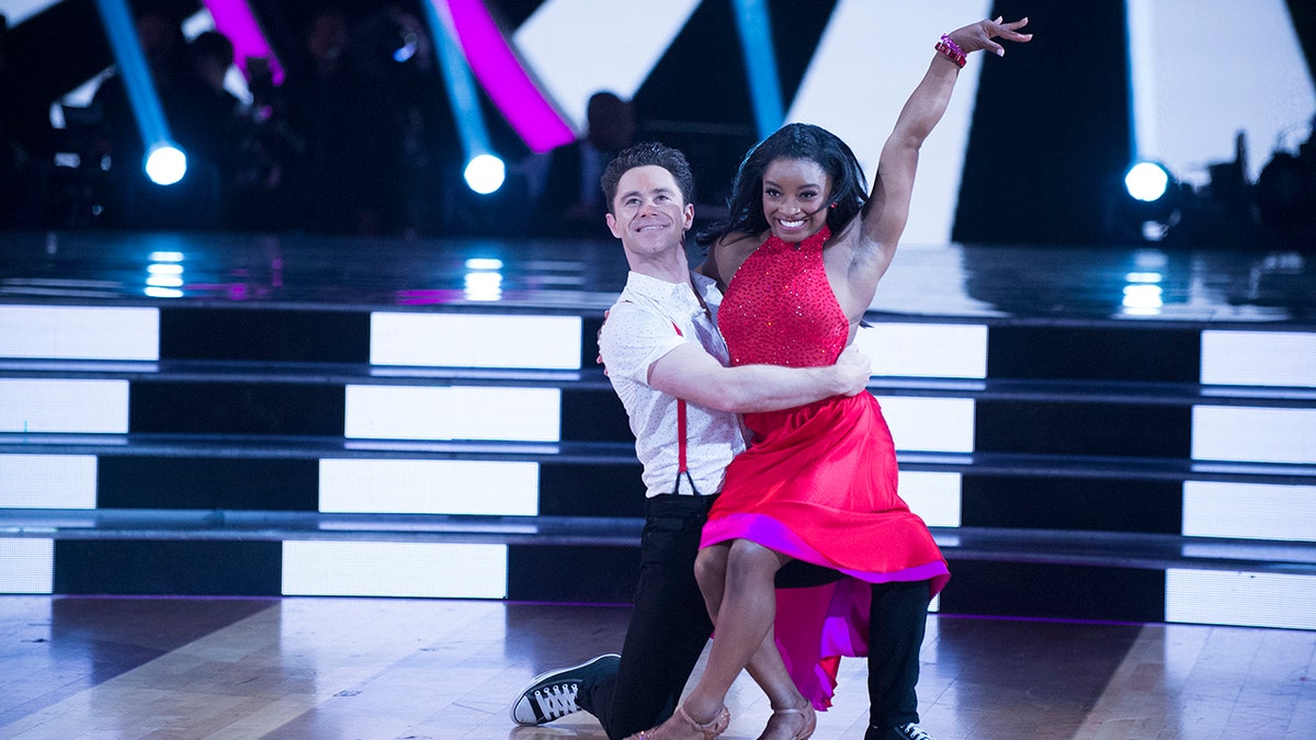 Sasha Farber in a white shirt knees as Simone Biles in a red dress puts her arm in the air and sits on him during 'Dancing with the Stars'