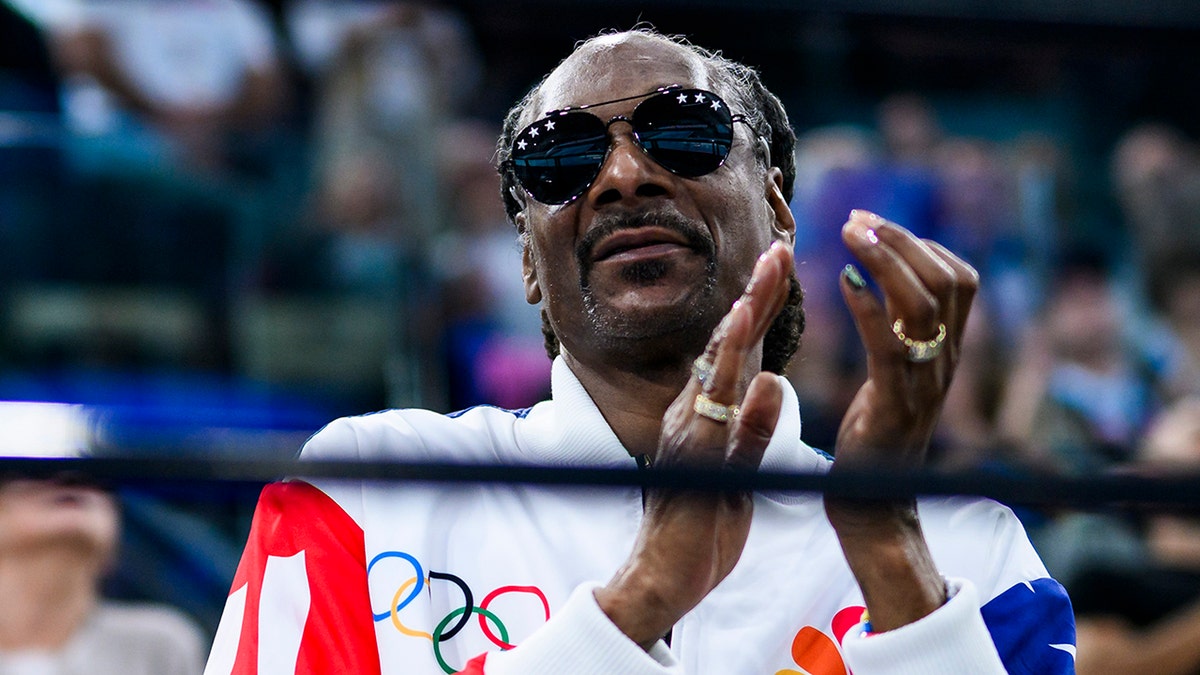 Snoop Dogg in a white Olympic jacket and sunglasses claps