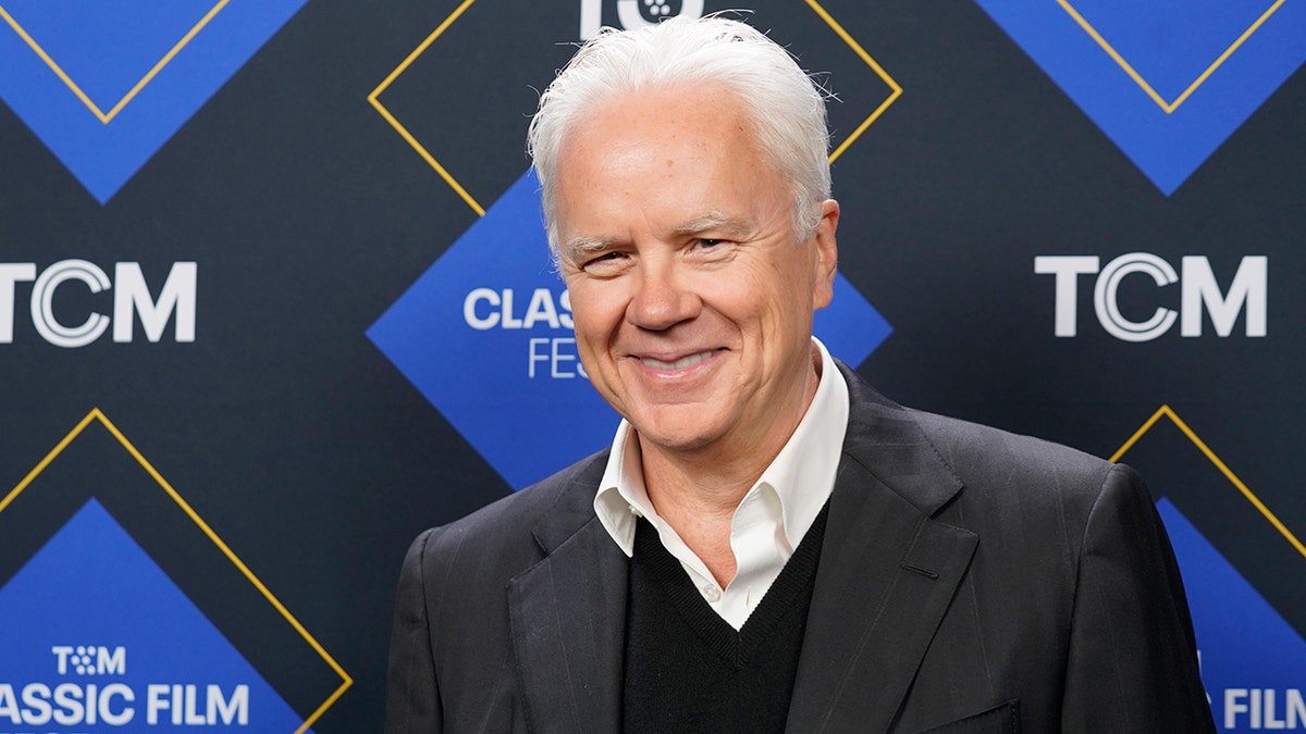 Tim Robbins in a grey suit and black sweater smiles for a photo