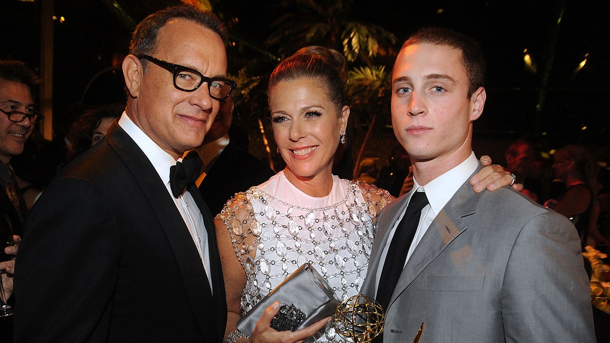 Tom Hanks con esmoquin y gafas de montura negra sonríe a la cámara junto a su mujer Rita Wilson con vestido blanco junto a su hijo Chet con traje gris y corbata negra